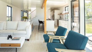 A modern living room with a white sofa, a coffee table, and blue armchairs. There is a kitchen area with a dining table and chairs visible in the background.
