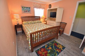 A bedroom with a wooden bed, a television on a cabinet, a nightstand with a lamp, and a floral rug. - Water's Edge Resort #210 - Oceanfront in Garden City Beach, SC (Garden City)