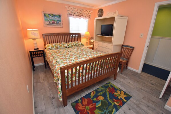 A bedroom with a wooden bed, a television on a cabinet, a nightstand with a lamp, and a floral rug. - Water's Edge Resort #210 - Oceanfront in Garden City Beach, SC (Garden City)