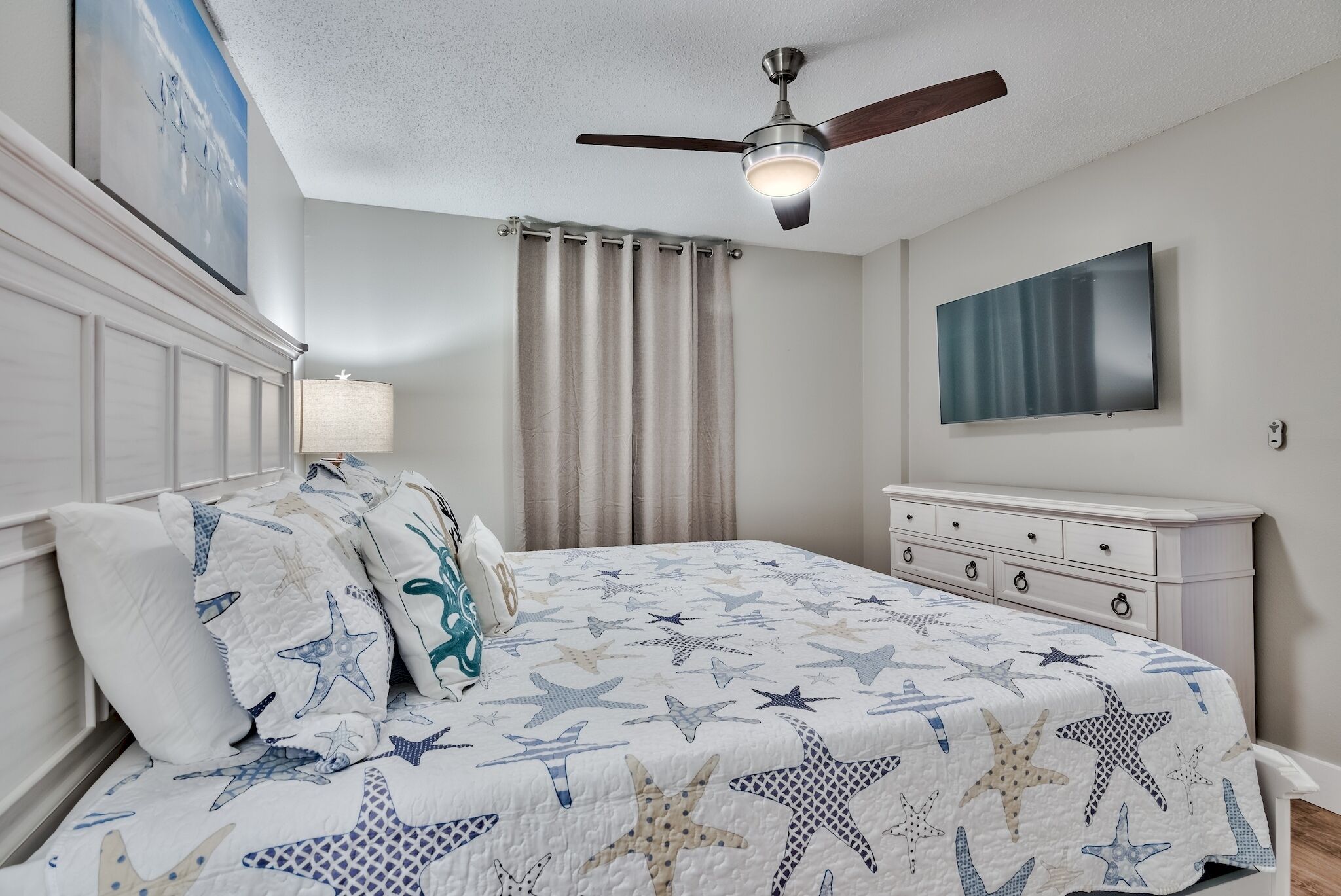 Un dormitorio con cama, ropa de cama con estampado de estrellas, ventilador de techo, televisor y mesita de noche.