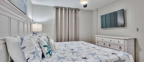 Un dormitorio con cama, ropa de cama con estampado de estrellas, ventilador de techo, televisor y mesita de noche.