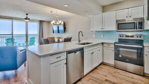 A modern kitchen with white cabinets, a dishwasher, a microwave, and an oven. There is a large window with a view of the ocean. - Gulf Front, Renovated unit in Pelican Beach Resort, Wi-Fi, Cable (Destin)