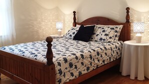 Un lit à baldaquin en bois, recouvert d’une couvre-lit à motifs floraux, deux tables de chevet avec des lampes et une fenêtre ornée de rideaux légers.