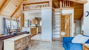 Interior acogedor de una cabaña con comedor de madera, cocina, sofá azul y habitación superior con escalera.