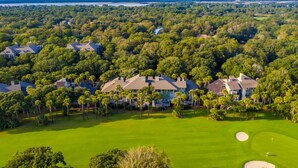 An aerial view of a golf course with multiple houses surrounded by dense trees and a clear view of the water body in the background.