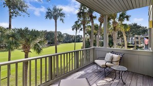 A covered balcony with a view of a golf course, a chair, and a small table.