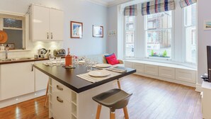 Una cocina moderna con armarios blancos, una isla central con taburetes, una mesa de comedor para cuatro personas y un ventanal con cortinas a rayas.