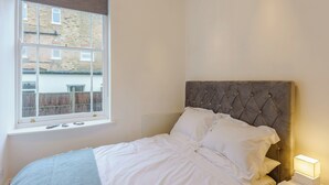 Una cama bien tendida con un cabecero gris, una lámpara de noche y una ventana con vista a un edificio de ladrillo.