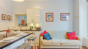 Una cocina moderna con armarios blancos, un comedor con una mesa para dos personas y un sofá con cojines de colores.