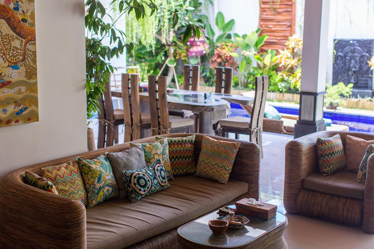 A cozy living area with a brown sofa, colorful cushions, a glass coffee table, and a view of a garden with plants and a fountain.