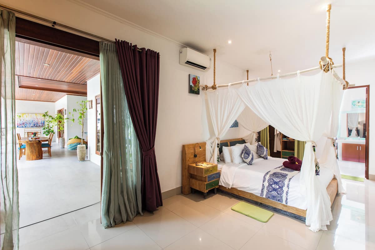 A bedroom with a four-poster bed, a bedside table, a green mat, and a view of the dining area.