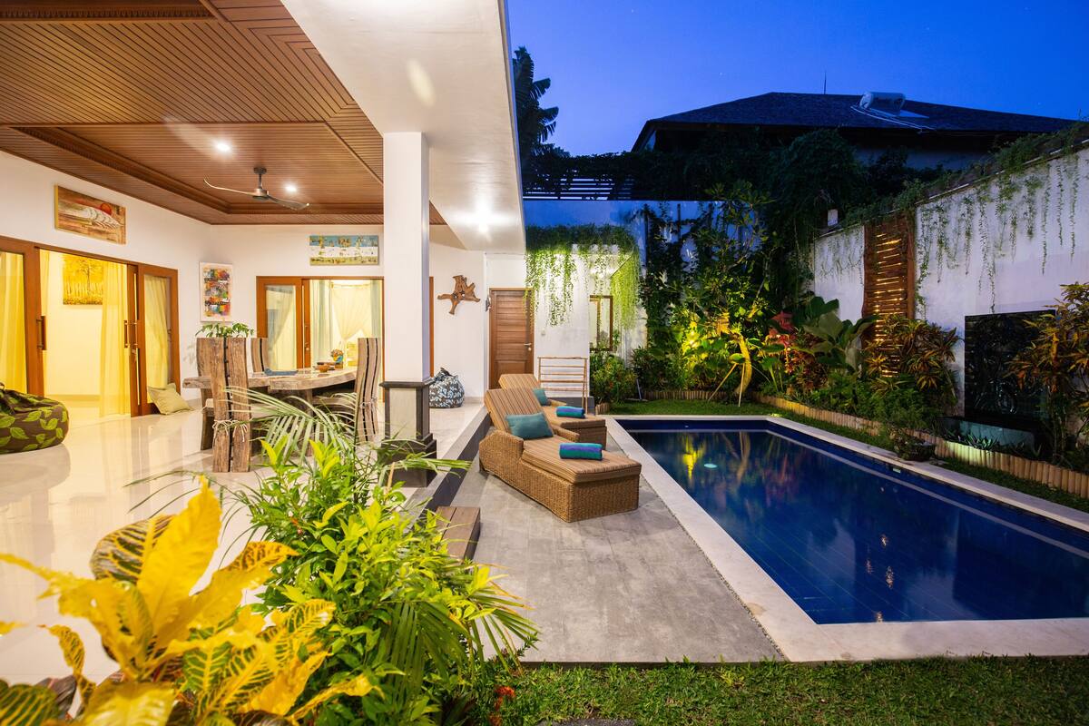 A modern house with a swimming pool, a patio with furniture, and lush greenery.
