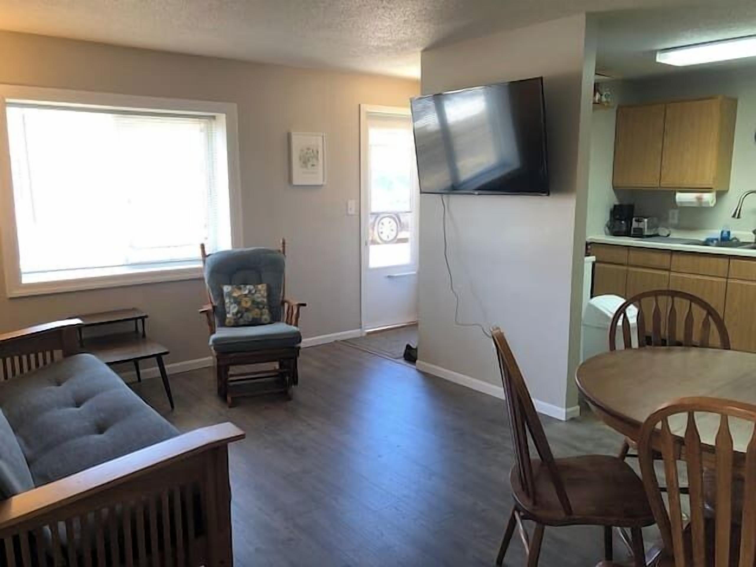 A living room with a sofa, a dining table, and chairs, a kitchen area with cabinets and a TV mounted on the wall.