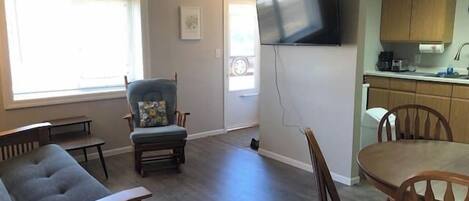 A living room with a sofa, a dining table, and chairs, a kitchen area with cabinets and a TV mounted on the wall.