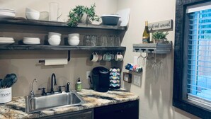 A kitchen with a granite countertop, stainless steel sink, and various kitchen utensils. There are shelves above the countertop holding dishes, glasses, and a potted plant. A sign on the wall reads "Coffee with a smile." - Secluded Peaceful Chalet 1 Bdrm King Suite AC, Private Laundry Room (Bigfork)