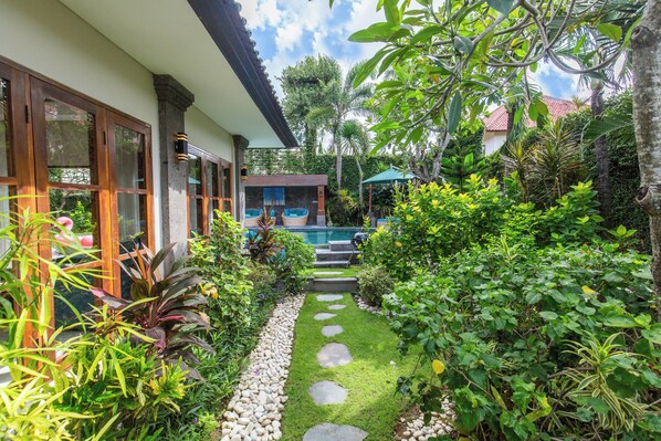 Un jardin luxuriant avec un sentier en pierre menant à une zone de piscine, entouré de plantes tropicales et bordé d’un bâtiment doté de grandes fenêtres.
