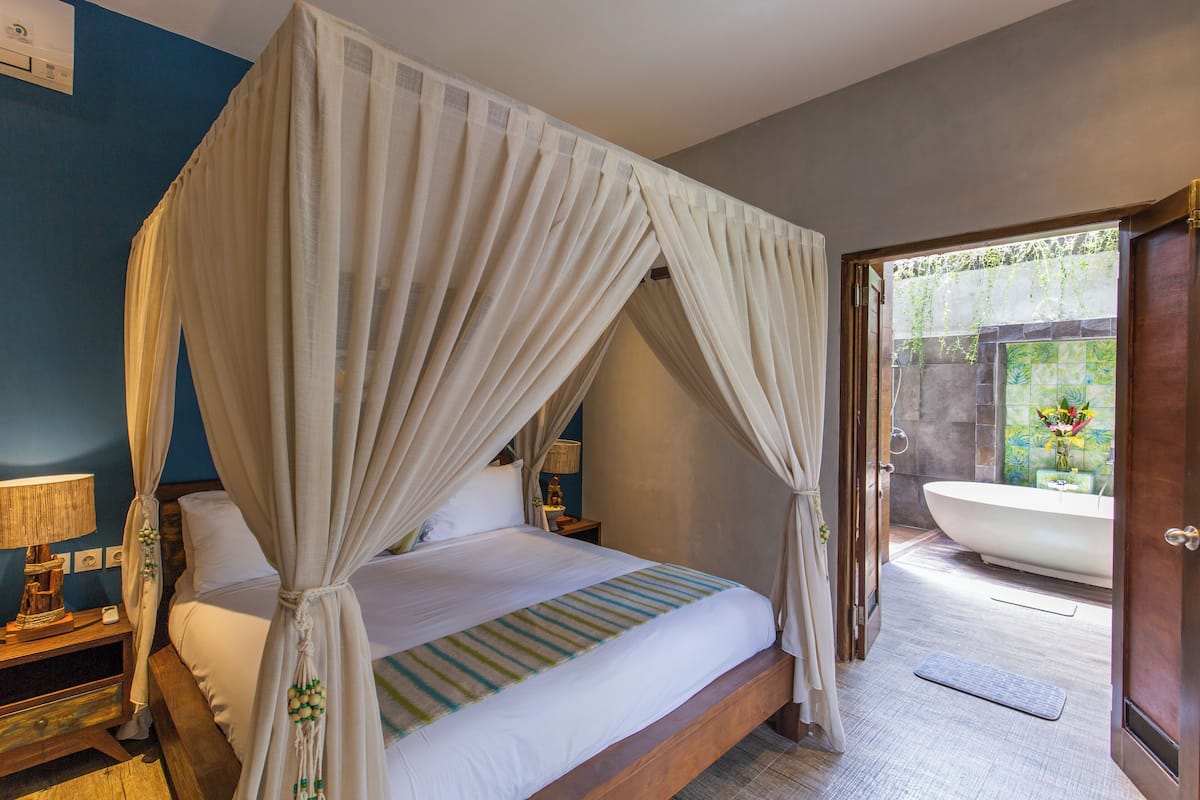 Un lit à baldaquin à quatre poteaux avec une structure en bois, une table de chevet avec une lampe, et une vue sur une salle de bain équipée d’une baignoire et d’une douche.