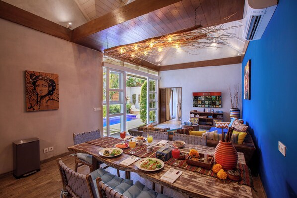 A dining area with a wooden table set for a meal, a sofa with cushions, and a large window with a view of greenery. - Delight ART Villas (Seminyak)