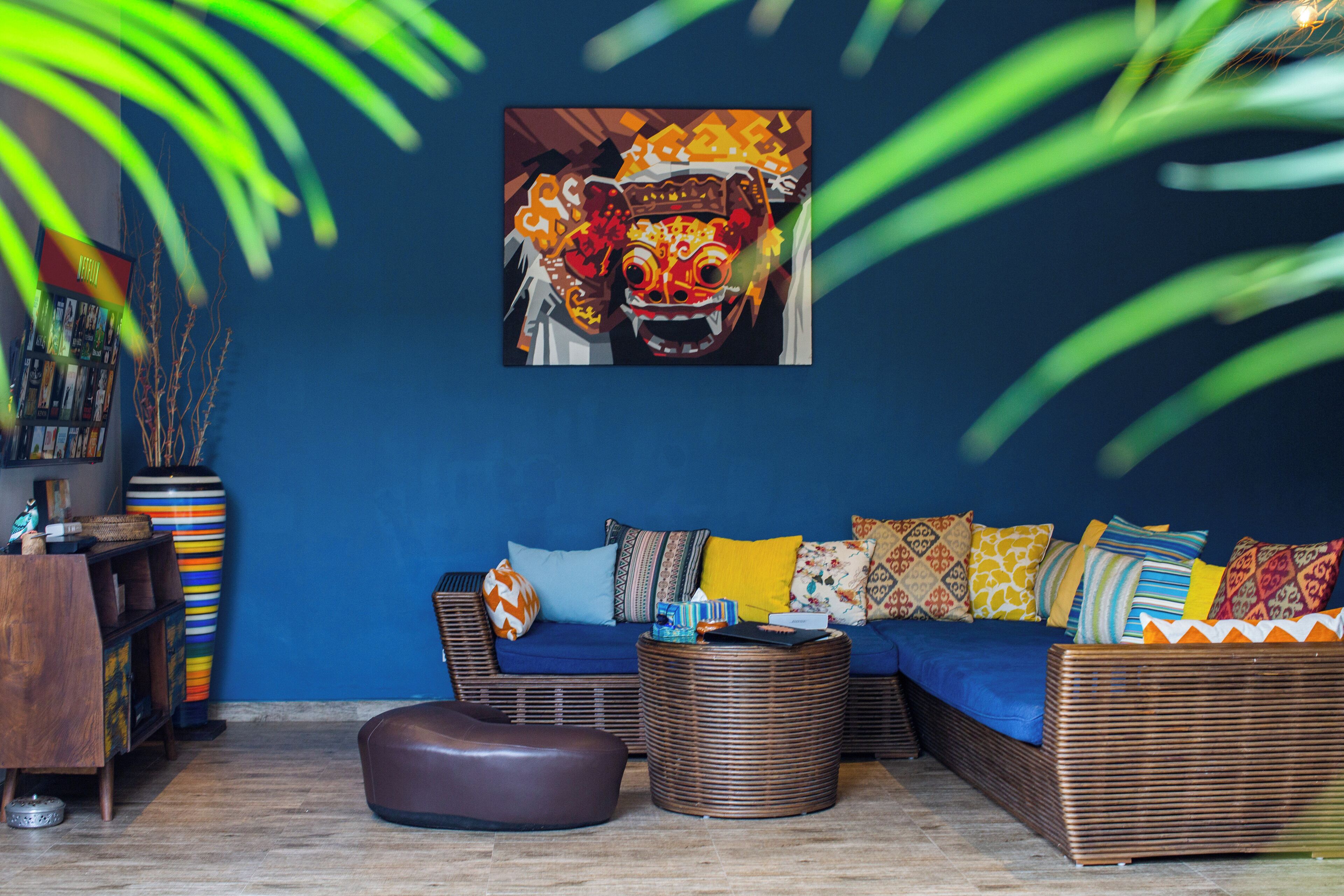 A room with a blue wall, a wicker sofa with colorful cushions, a brown ottoman, and a large painting of a traditional mask.