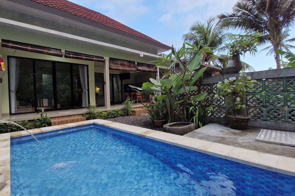 Uma piscina com uma estátua de pedra, uma casa com telhado de cerâmica e plantas tropicais.