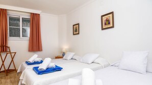 A hotel room with two beds, white linens, blue towels, a window with curtains, a small table with a lamp, and two framed pictures on the wall.