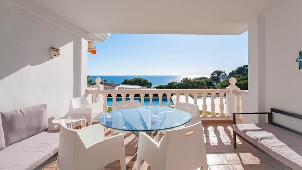 A balcony with a glass table and chairs, a sofa, and a view of the sea and greenery.