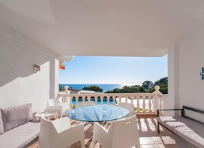 A balcony with a glass table and chairs, a sofa, and a view of the sea and greenery.