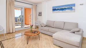 A living room with a beige sofa, a wooden coffee table with books and a vase, a white chair, and a glass door leading to an outdoor area. - Poolbeach VI 4 pax - Moraira, Costa Blanca (Moraira)