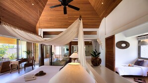 A spacious bedroom with a large bed, a wooden ceiling fan, a bedside table, a lamp, and a view of the outdoors. - Baan Kilee (Koh Samui)