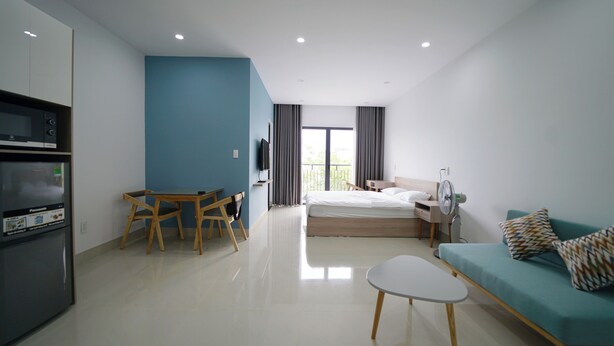 A modern hotel room with a bed, a sofa, a dining table with chairs, a kitchenette, and a large window with curtains. - Sunny Studio with balcony ★ Steps to Dragon Bridge (Đà Nẵng)