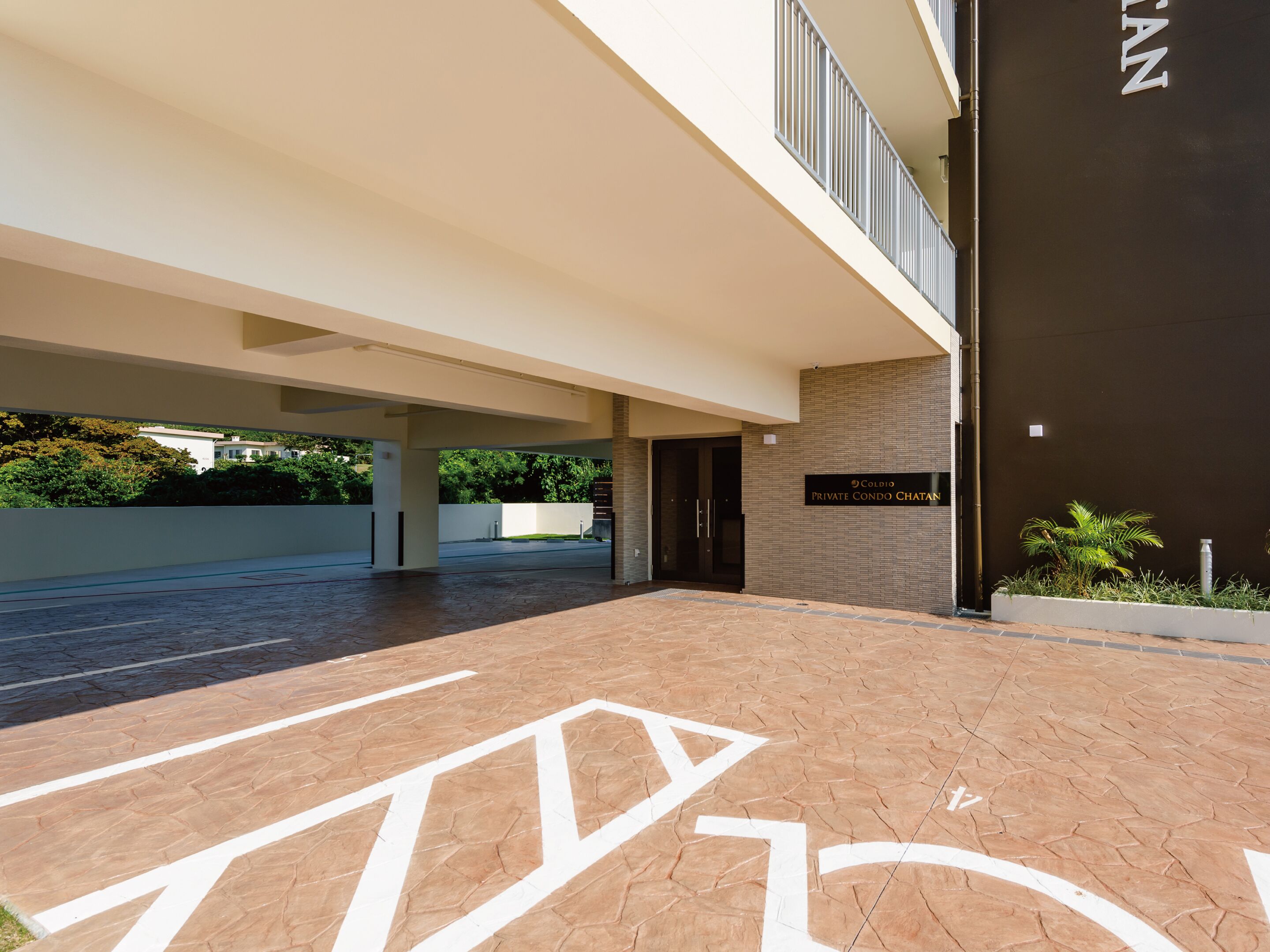 Entrada moderna de un edificio con una galería techada, un cartel que dice 'Coldio Private Condo Chatan' y un aparcamiento para bicicletas.