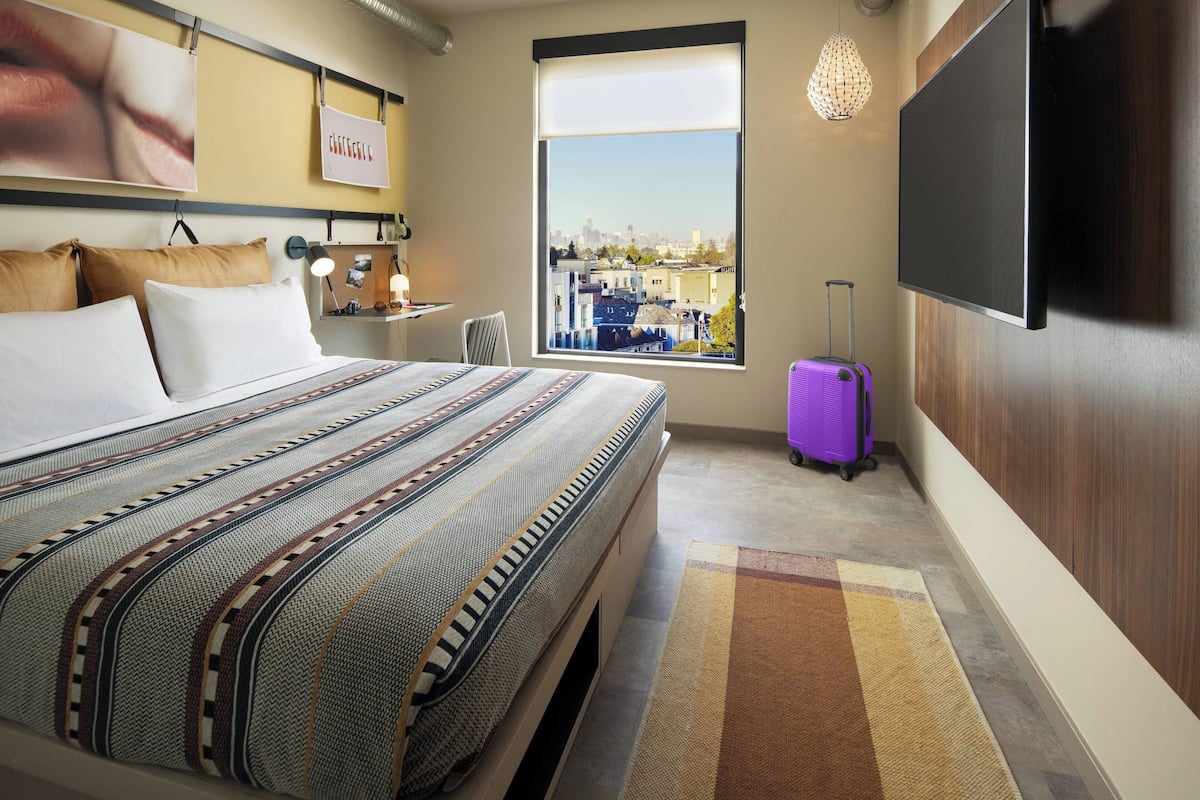 A hotel room with a large bed, a city view window, a flat-screen TV, and a purple suitcase.