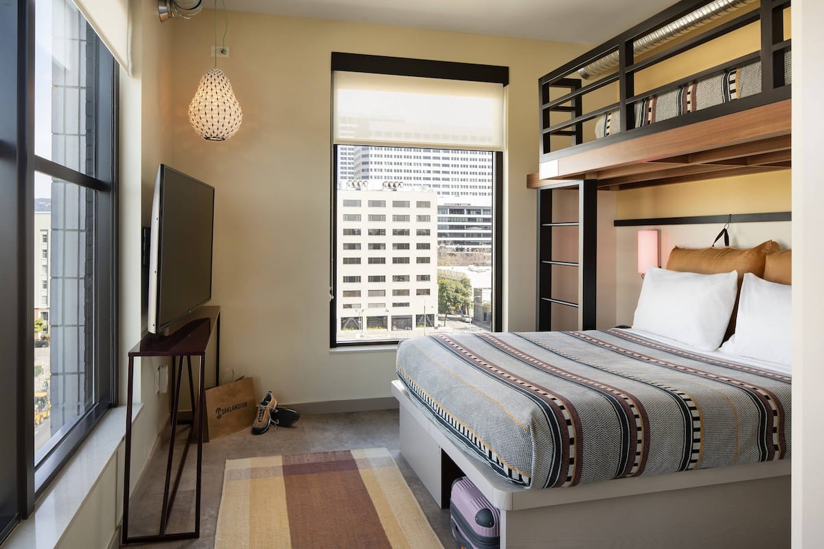 A bedroom with a bunk bed, a television on a stand, a window with a view of buildings, and a patterned rug.
