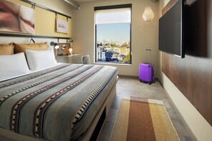 Habitació d'hotel amb un llit gran, una finestra amb vista a la ciutat, un televisió de pantalla plana i una maleta morada.