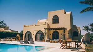 Zona piscina con lettini e tavolo da pranzo, circondata da un edificio con finestre ad arco e tetto in tegole.