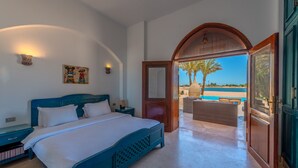 Una camera da letto con un letto, comodini, una lampada a parete e vista su una spiaggia e palme attraverso una porta aperta.