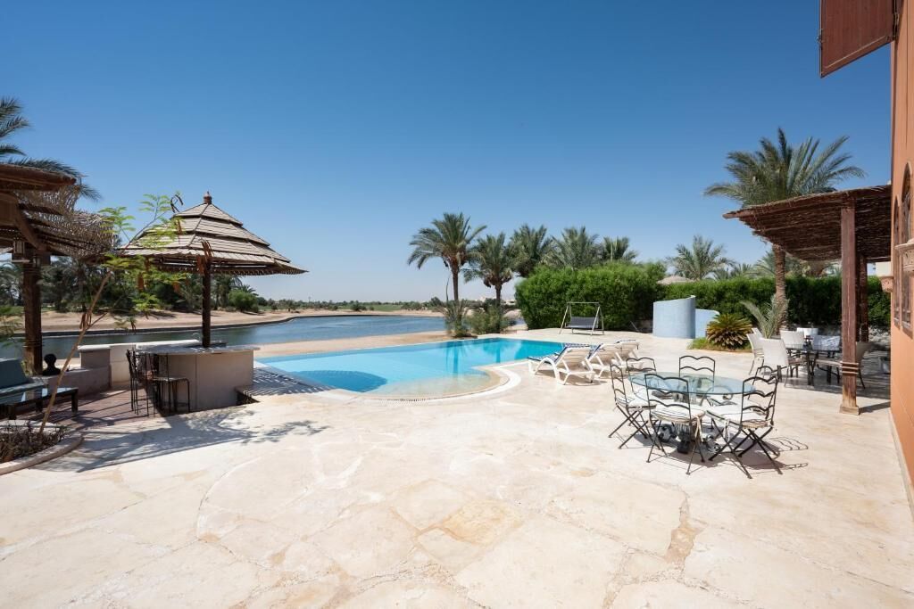 A pool area with lounge chairs and a table, surrounded by palm trees and a clear sky.