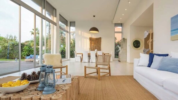 Un salon moderne doté de grandes fenêtres, d’un canapé blanc, de chaises en bois et d’un coin repas.