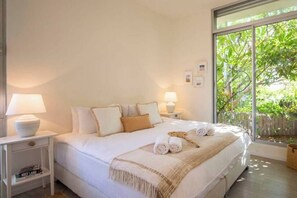 A bedroom with a bed, bedside lamps, a nightstand with books, and a view of greenery outside.