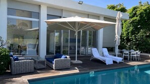 Un espace aménagé au bord de la piscine, avec des chaises longues, un coin salon abrité et une véranda fermée par des baies vitrées.