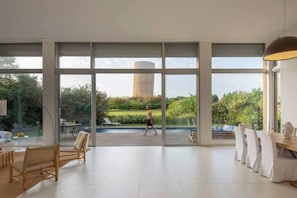 A spacious room with large windows, a dining area, and a view of a garden and a tall structure outside.