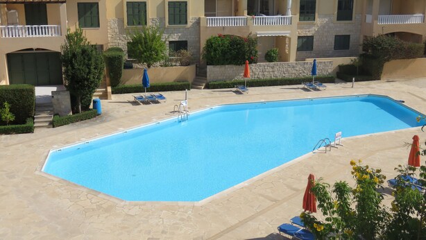 A rectangular swimming pool with lounge chairs and umbrellas, surrounded by stone-paved walkways and a building with balconies and satellite dishes. - G3 Polis Gardens - includes WiFi & A/C  (Polis)