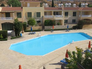 Une piscine rectangulaire, agrémentée de chaises longues et de parasols, entourée de chemins pavés de pierres et d’un bâtiment doté de balcons et d’antennes paraboliques.