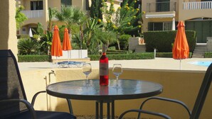 A round table with two chairs, a bottle of red wine, and two wine glasses on a balcony overlooking a swimming pool and a landscaped garden. - G3 Polis Gardens - includes WiFi & A/C  (Polis)