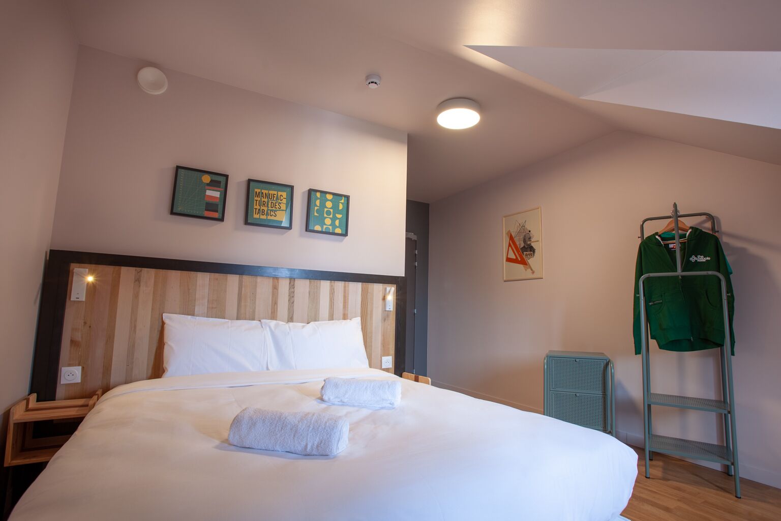 Una cama bien hecha con sábanas blancas y dos toallas, un cabecero de madera y una chaqueta verde en un perchero.