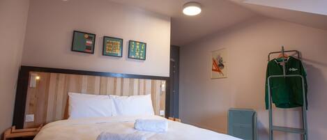 Una cama bien hecha con sábanas blancas y dos toallas, un cabecero de madera y una chaqueta verde en un perchero.