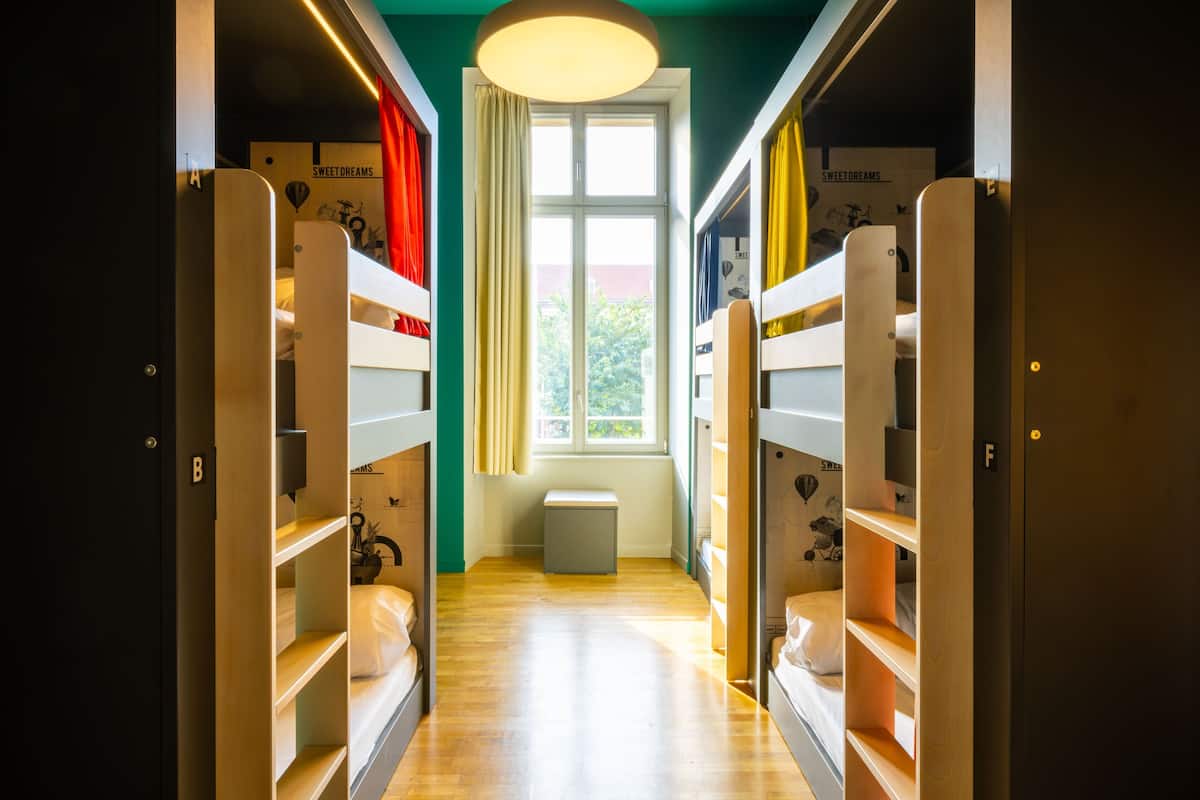 Ein schmaler Gang mit Etagenbetten an beiden Seiten, ein kleines Fenster mit Blick auf Grünflächen und eine Leuchte darüber.