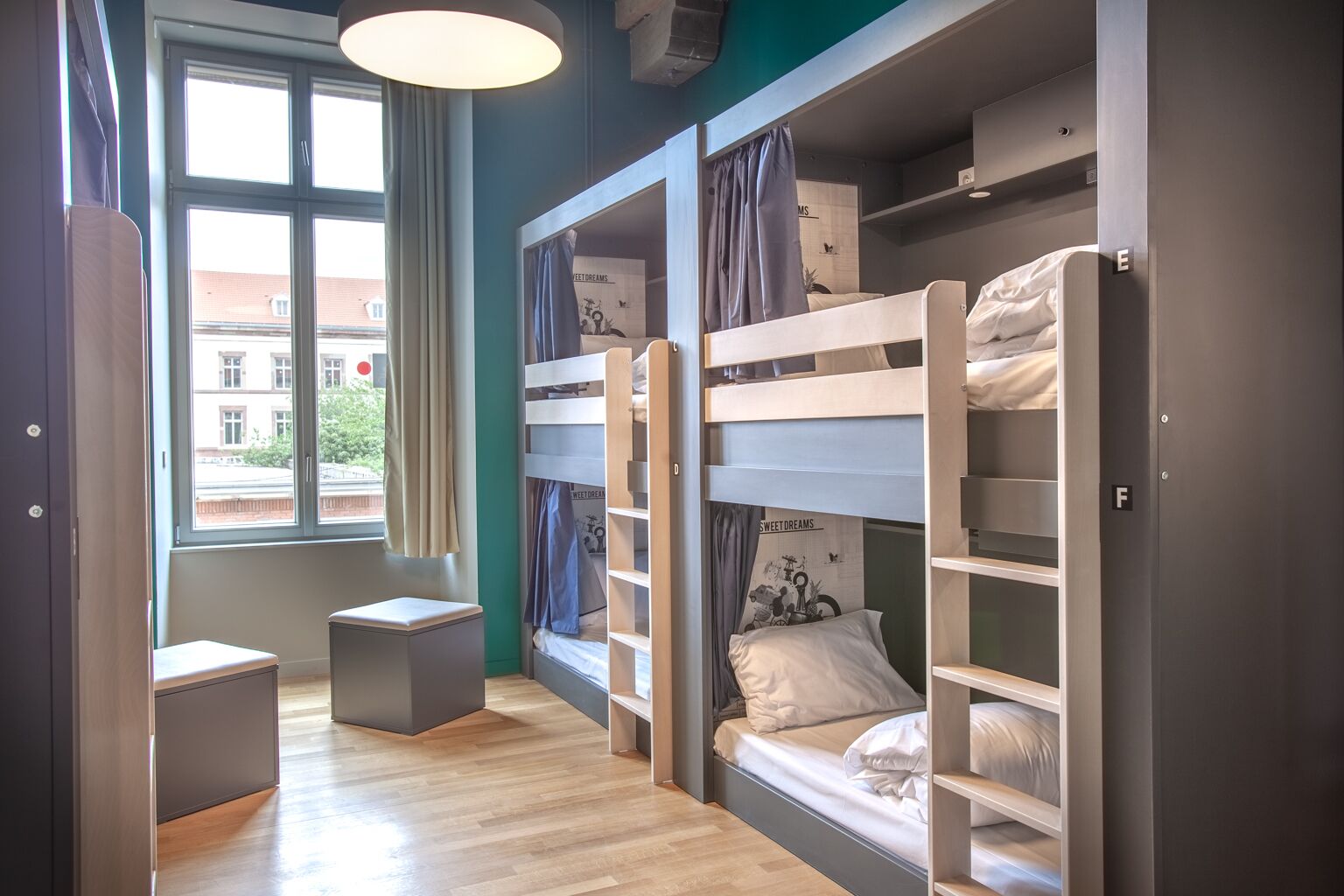 Een slaapkamer met stapelbed, een raam, een klein tafeltje en een deur met de aanduiding 'F'.