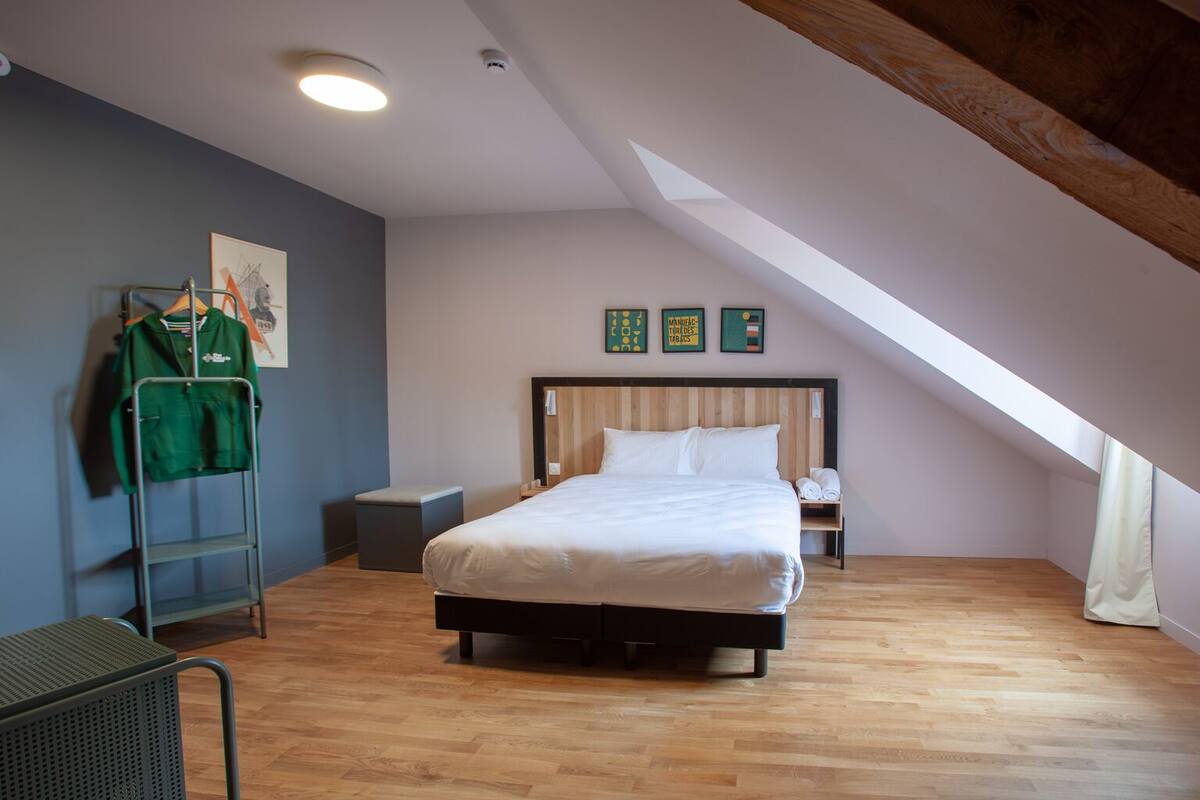 Ein Schlafzimmer mit einem Bett, einem kleinen Nachttisch, einem Stuhl, einem Kleiderhaken mit Kleidung und einer Deckenleuchte.