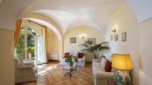Ein geräumiges Wohnzimmer mit einer weißen Sofagarnitur, einem Couchtisch und einer Lampe. Ein großes Fenster mit Vorhängen, ein Topfpflanze und Blick ins Grüne sind ebenfalls vorhanden.